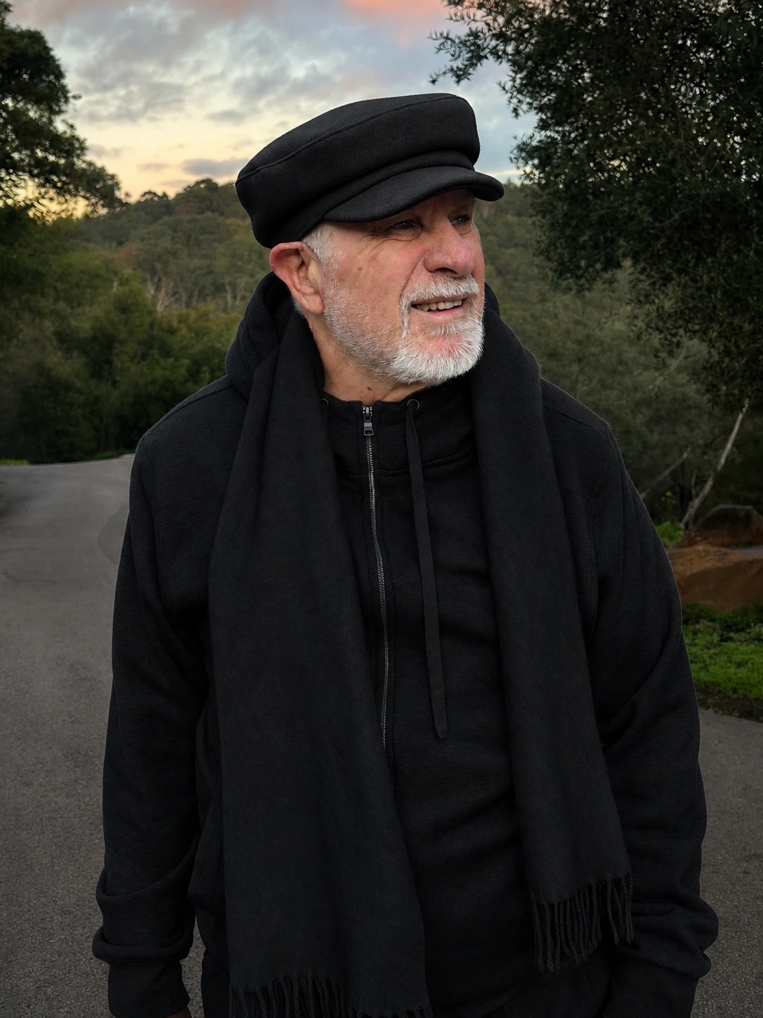 Vishrant outdoors in a black cap and scarf, smiling, trees behind him.